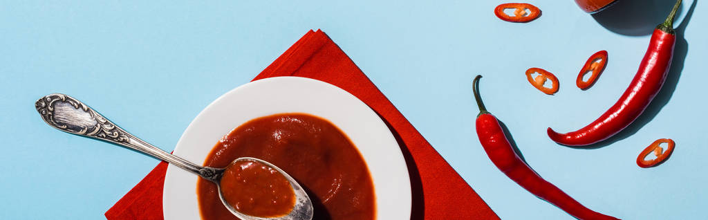 Верхний вид на вкусный пряный кетчуп в тарелке с спелыми перцами чили на голубой поверхности, панорамный снимок
 - Фото, изображение