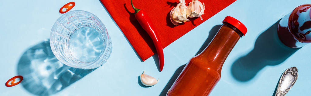 Верхний вид бутылок с кетчупом, чеснок с перцем чили и стакан воды на синей поверхности, панорамный снимок
 - Фото, изображение
