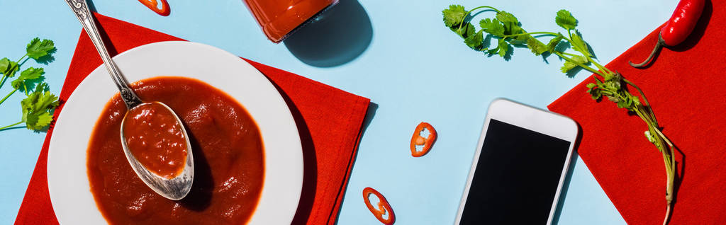 Bovenaanzicht van ketchup met chili peper en koriander naast smartphone met blanco scherm op blauw oppervlak, panoramisch shot - Foto, afbeelding