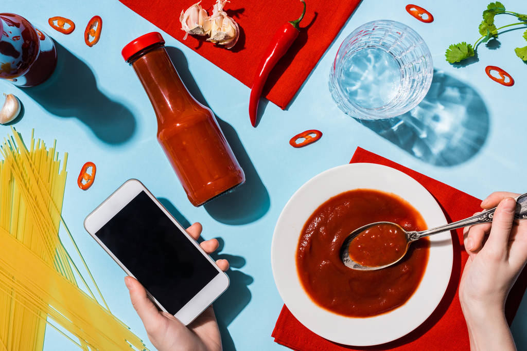 Vista recortada de la mujer sosteniendo el teléfono inteligente junto a ketchup, espaguetis y vaso de agua en la superficie azul
 - Foto, Imagen