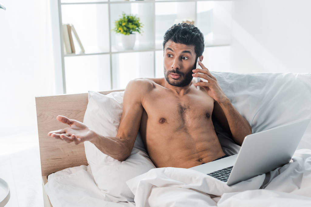 bonito e chocado bi-racial homem com laptop falando no smartphone na manhã
  - Foto, Imagem