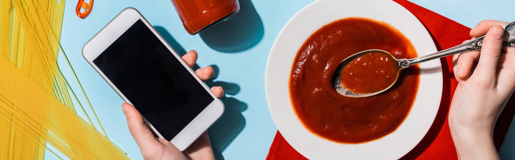 Vista cortada de mulher segurando smartphone ao lado de ketchup e espaguete no fundo azul, tiro panorâmico
 - Foto, Imagem