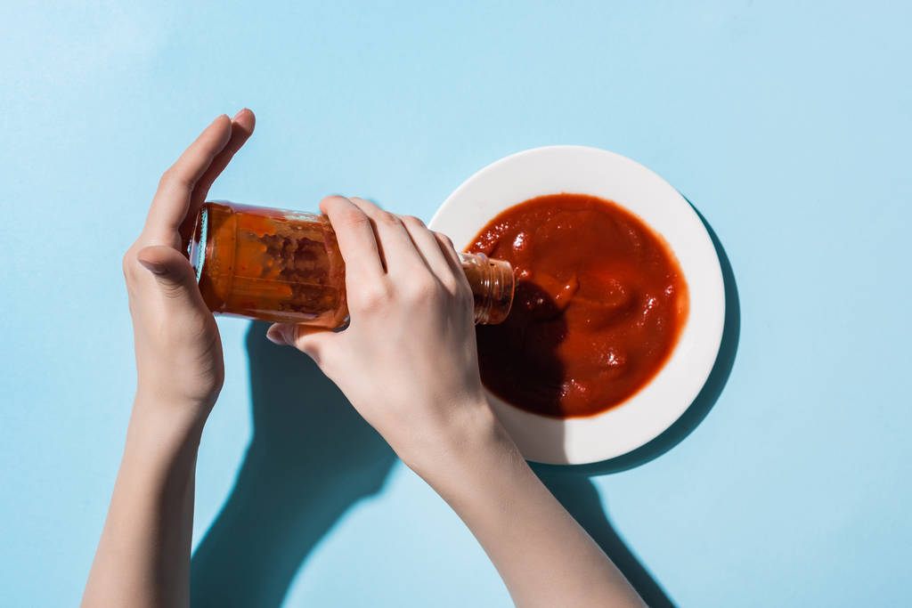 Обрезанный вид женщины, наливающей томатный соус из бутылки в тарелку на синем фоне
 - Фото, изображение