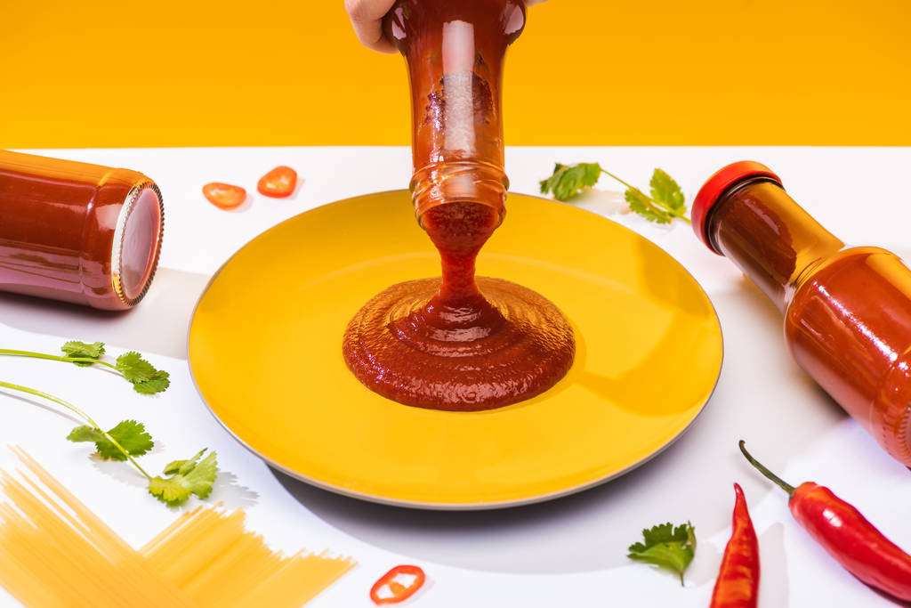 Vista recortada de la mujer vertiendo ketchup en el plato junto a espaguetis y chiles en la superficie blanca aislada en amarillo
 - Foto, Imagen