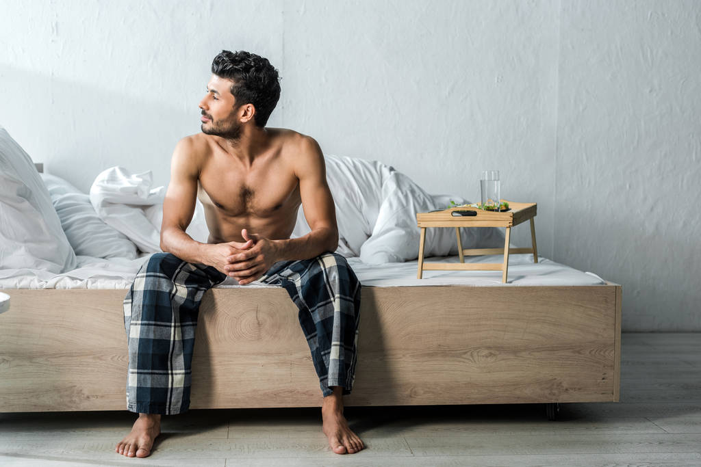 handsome bi-racial man sitting on bed and looking away in morning  - Photo, Image