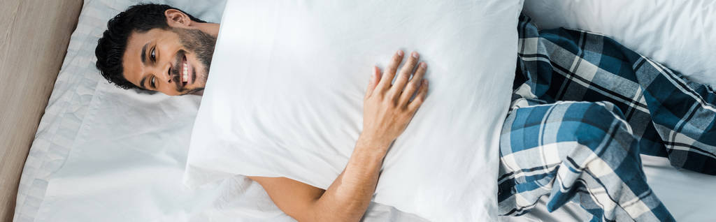 панорамный снимок красивого и улыбающегося двурасового мужчины, лежащего на кровати с подушкой утром
  - Фото, изображение