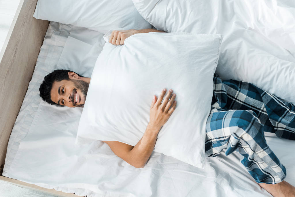 vista superior del hombre bi-racial guapo y sonriente acostado en la cama con almohada en la mañana
  - Foto, Imagen