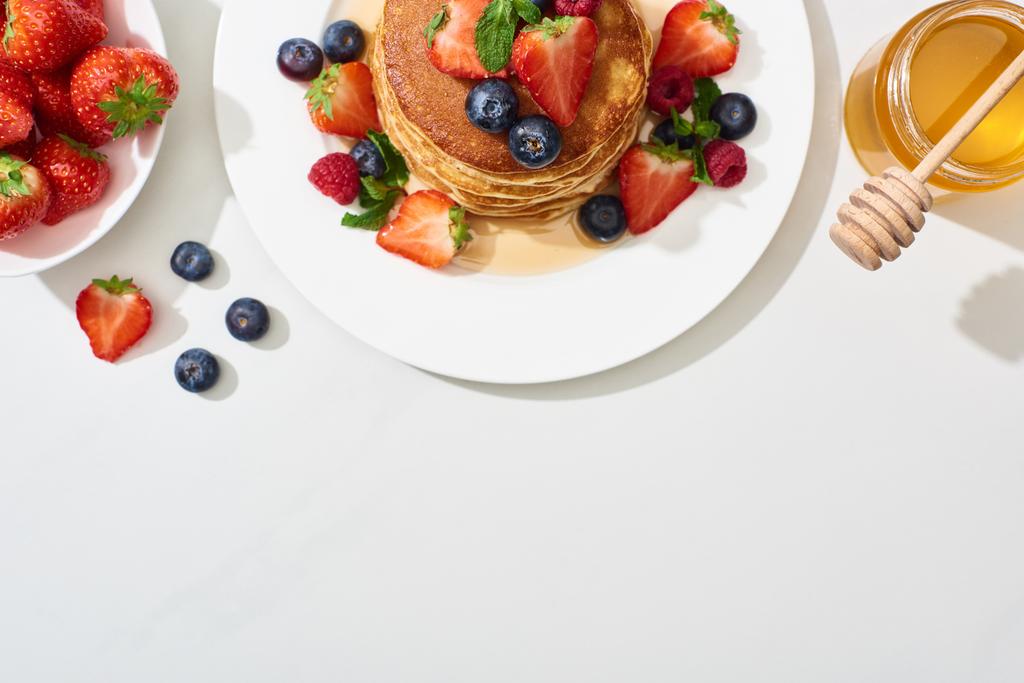 bovenaanzicht van heerlijke pannenkoeken met honing, bosbessen en aardbeien op bord op marmeren wit oppervlak - Foto, afbeelding