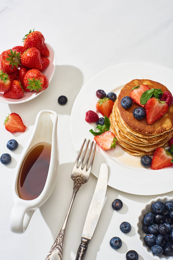 vue de dessus de délicieuses crêpes au sirop d'érable, bleuets et fraises sur assiette près de la fourchette et couteau sur surface marbre blanc
 - Photo, image