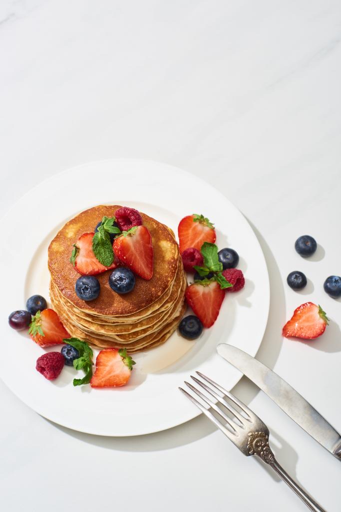 Draufsicht auf leckere Pfannkuchen mit Ahornsirup, Blaubeeren und Erdbeeren auf Teller in der Nähe von Gabel und Messer auf marmorweißer Oberfläche - Foto, Bild