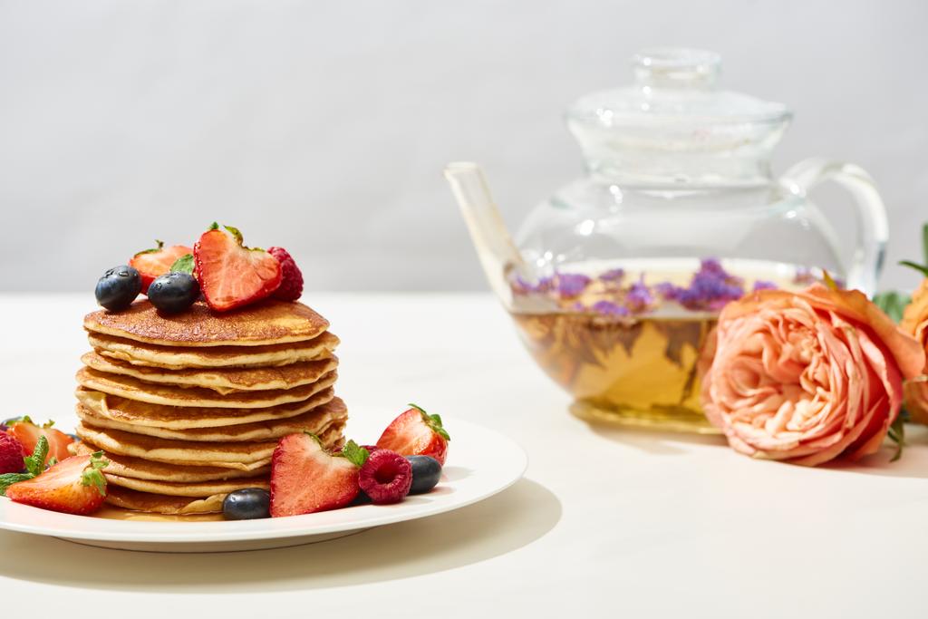 επιλεκτική εστίαση των νόστιμα τηγανίτες με βατόμουρα και φράουλες στο πιάτο κοντά τριαντάφυλλο λουλούδια και τσάι από βότανα στη λευκή επιφάνεια απομονώνονται σε γκρι - Φωτογραφία, εικόνα