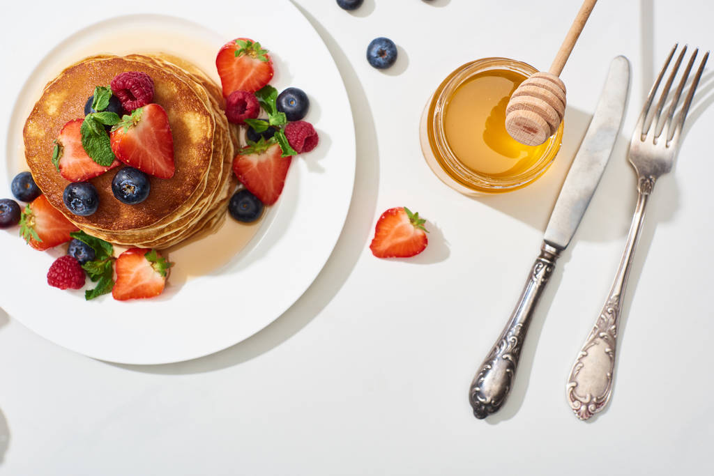 vista superior de deliciosos panqueques con miel, arándanos y fresas en el plato cerca de cubiertos en la superficie blanca de mármol
 - Foto, imagen