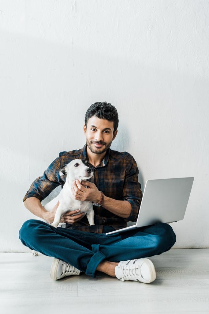 όμορφος και χαμογελαστός bi-φυλετικός άντρας με φορητό υπολογιστή κρατώντας Jack Russell Terrier - Φωτογραφία, εικόνα