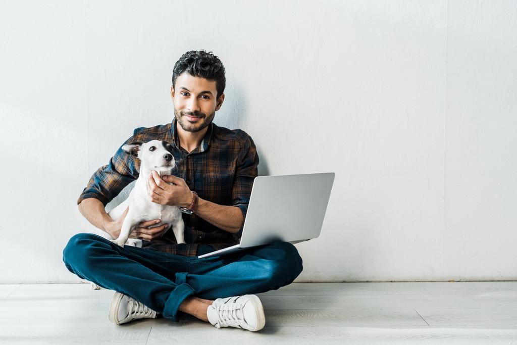 przystojny i uśmiechnięty birasowy mężczyzna z laptop gospodarstwa Jack Russell terrier - Zdjęcie, obraz