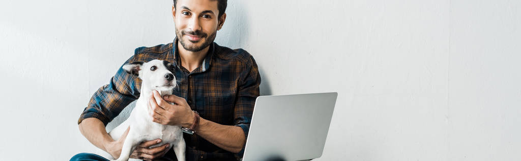 панорамный снимок красивого и улыбающегося бирасового человека с ноутбуком, держащего Джека Рассела терьера
 - Фото, изображение