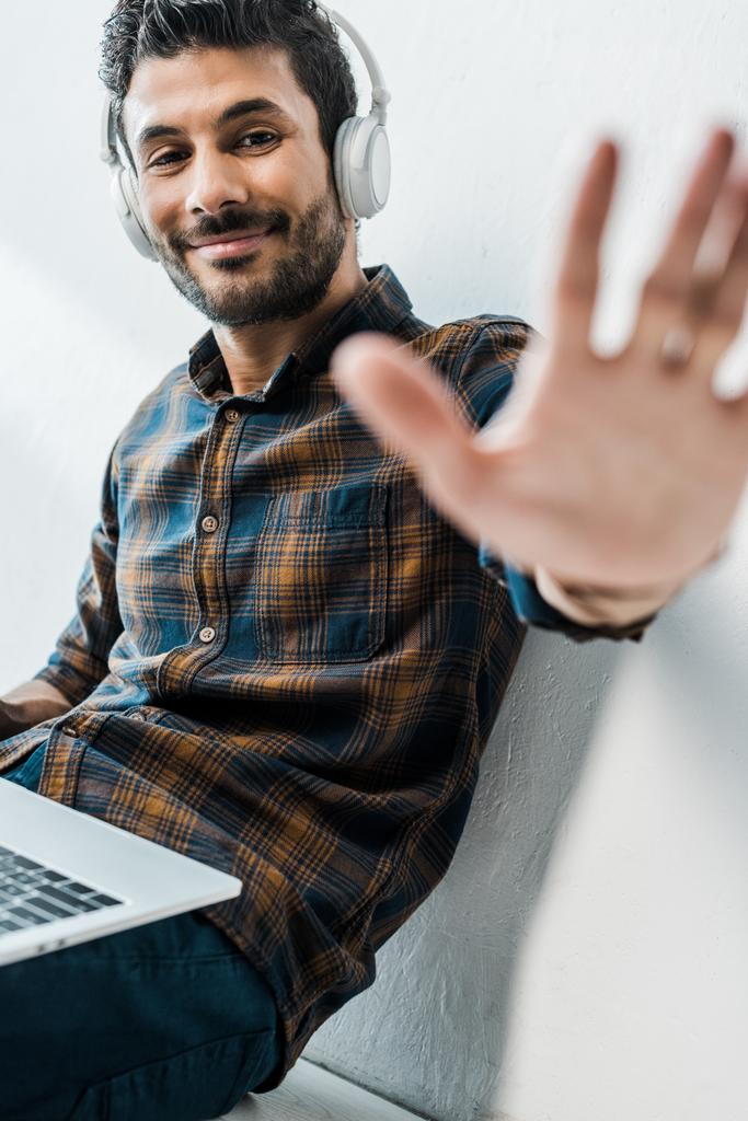 selective focus of handsome and smiling bi-racial man with laptop waving at camera  - Photo, Image