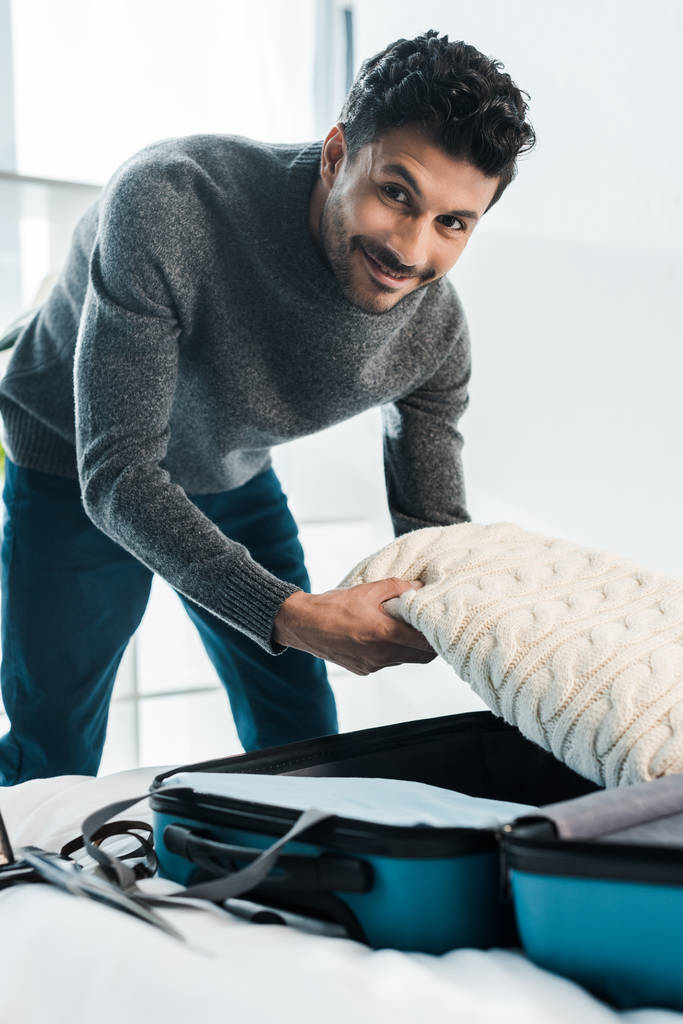 guapo y sonriente bi-racial hombre en suéter embalaje bolsa de viaje en apartamento
  - Foto, imagen