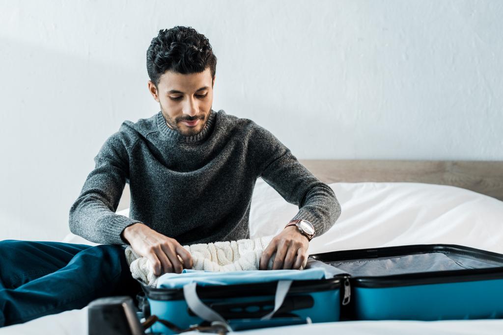 handsome bi-racial man in sweater packing travel bag in apartment  - Photo, Image
