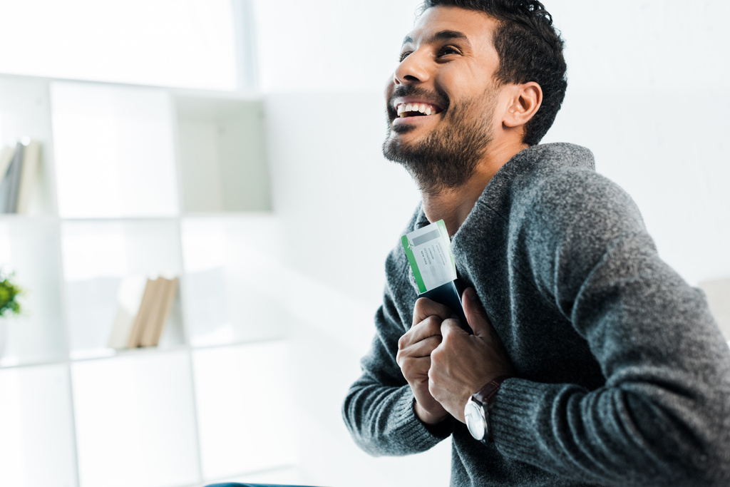 handsome and smiling bi-racial man in sweater holding passport with air ticket  - Photo, Image