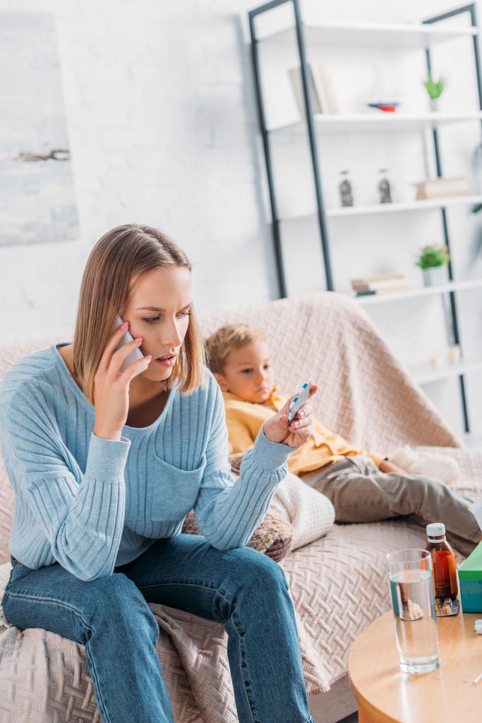 Besorgte Frau hält Tablettenblase in der Hand und spricht auf Smartphone, während sie neben krankem Sohn sitzt - Foto, Bild