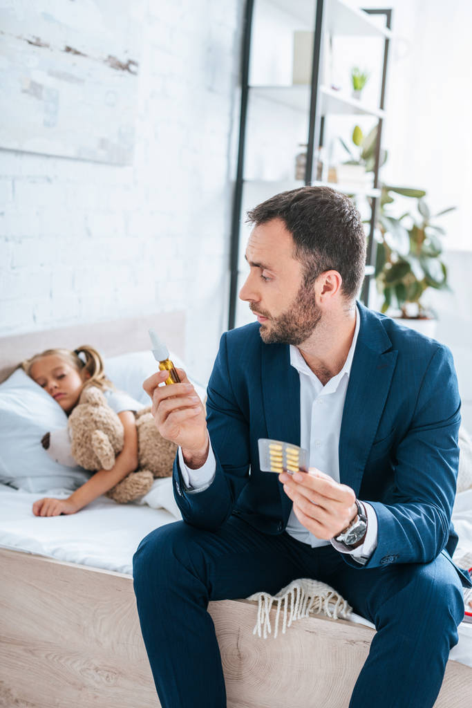 Vater hält Tabletten und Nasenspray in der Hand, während er neben kranker Tochter im Bett sitzt - Foto, Bild