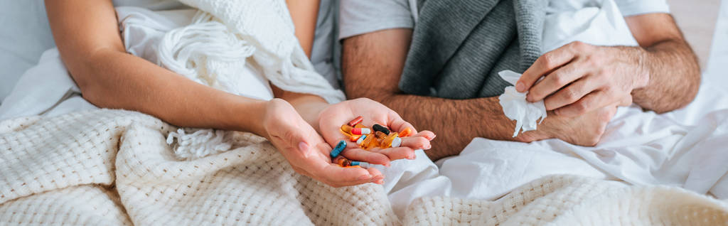 abgeschnittene Ansicht einer kranken Frau, die eine Handvoll Tabletten in der Hand hält, während sie neben ihrem kranken Mann im Bett liegt, Panoramaaufnahme - Foto, Bild