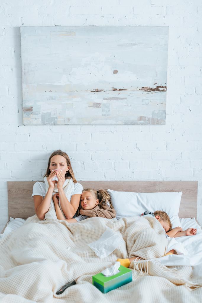 病気の女がナプキンでくしゃみをしながら病気の子供の近くのベッドに座って - 写真・画像