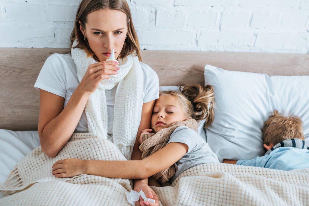 mulher doente usando spray nasal enquanto sentado na cama perto de crianças doentes
 - Foto, Imagem