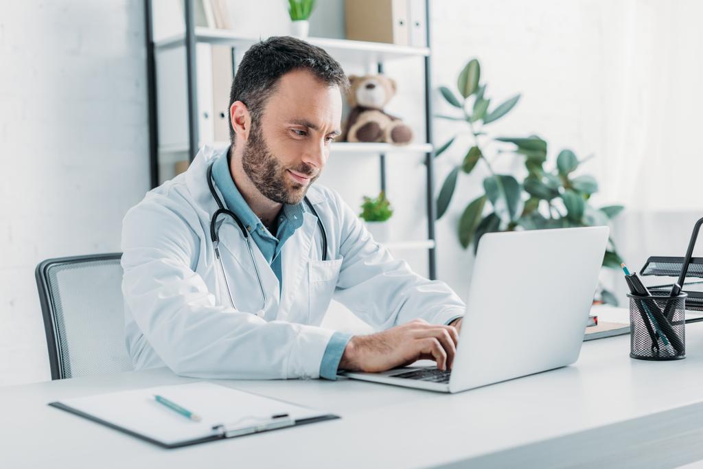pozitivní lékař sedí na pracovišti a pomocí notebooku - Fotografie, Obrázek