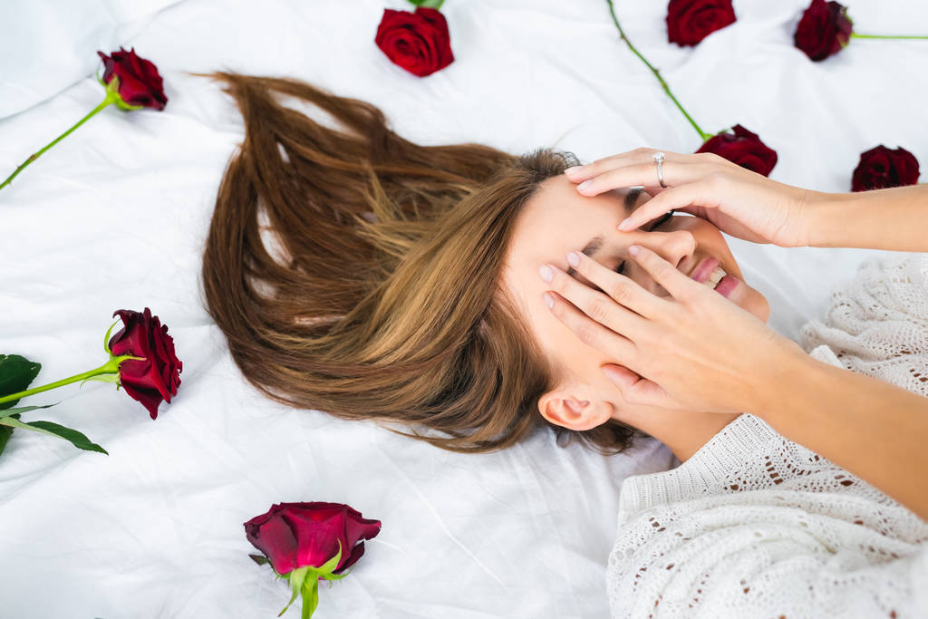 usmívající se žena zakrývající tvář a ležící na posteli s růžemi  - Fotografie, Obrázek