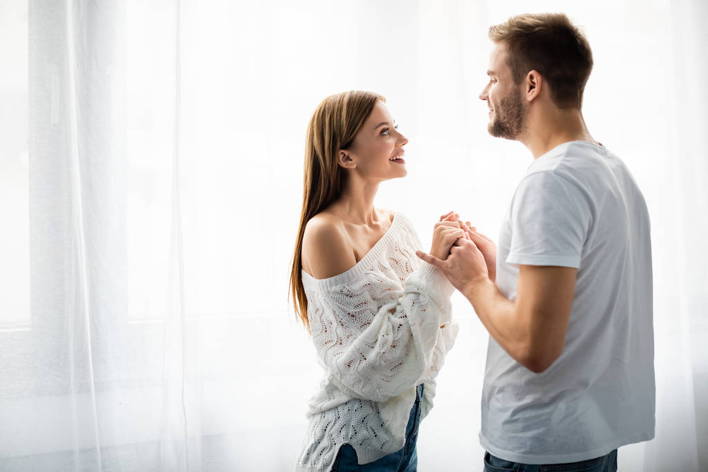 πλαϊνή άποψη του όμορφου άνδρα κρατώντας το χέρι με χαμογελαστή γυναίκα στο διαμέρισμα  - Φωτογραφία, εικόνα