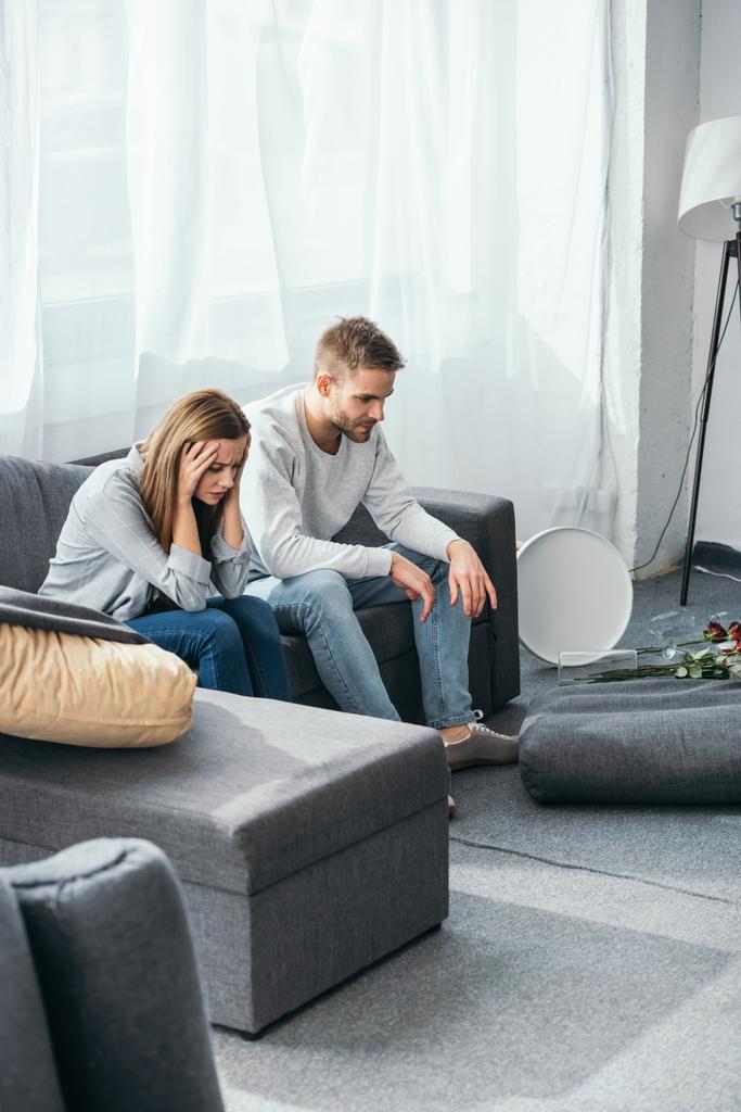 Traurige Frau und schöner Mann sitzen auf Sofa in beraubter Wohnung  - Foto, Bild