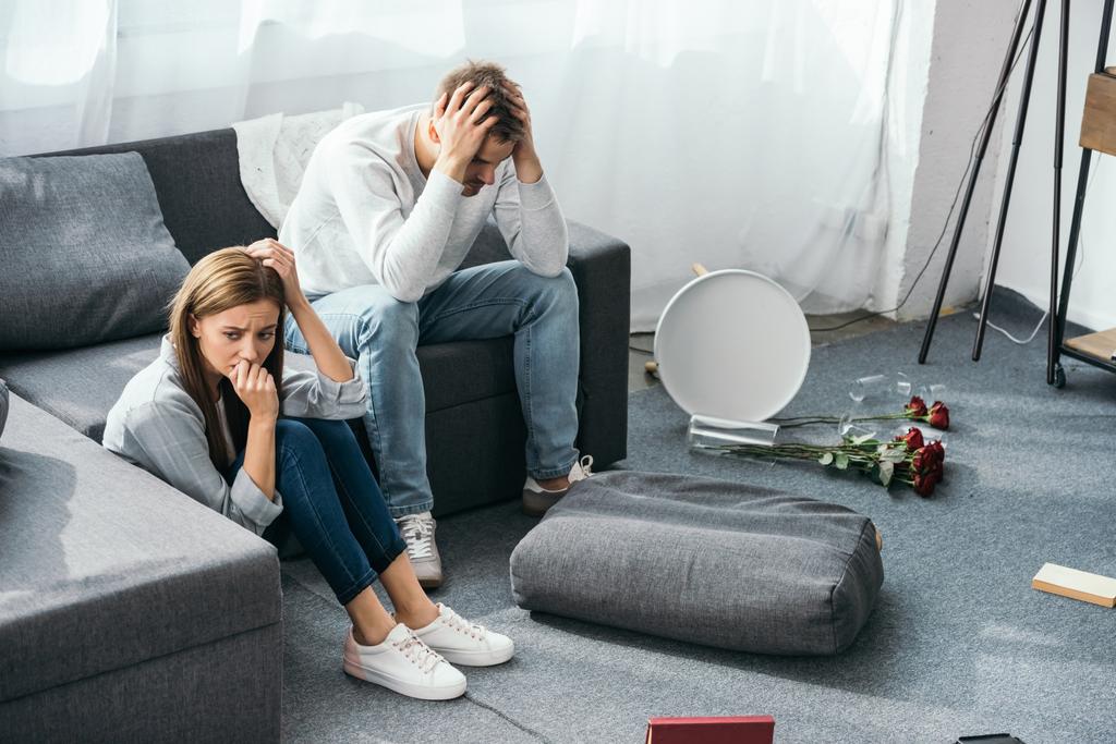 Traurige Frau am Boden und schöner Mann auf Sofa in beraubter Wohnung  - Foto, Bild