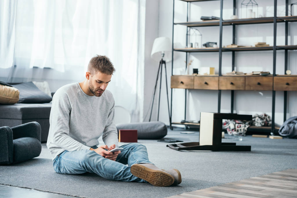 όμορφος άντρας κάθεται στο πάτωμα και χρησιμοποιεί smartphone σε διαμέρισμα ληστείας  - Φωτογραφία, εικόνα