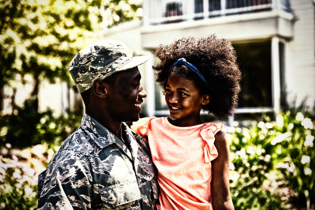 Vista frontal de perto de um jovem soldado afro-americano adulto no jardim do lado de fora de sua casa, segurando sua filha, ambos olhando um para o outro sorrindo
 - Foto, Imagem