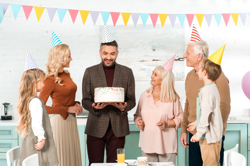 щасливий чоловік тримає торт на день народження, стоячи поруч з веселою сім'єю
 - Фото, зображення