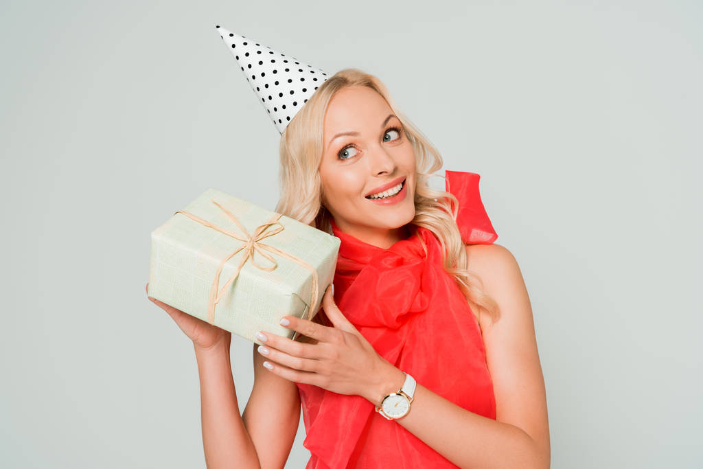 glimlachende, nieuwsgierige vrouw schudden geschenk doos geïsoleerd op grijs - Foto, afbeelding