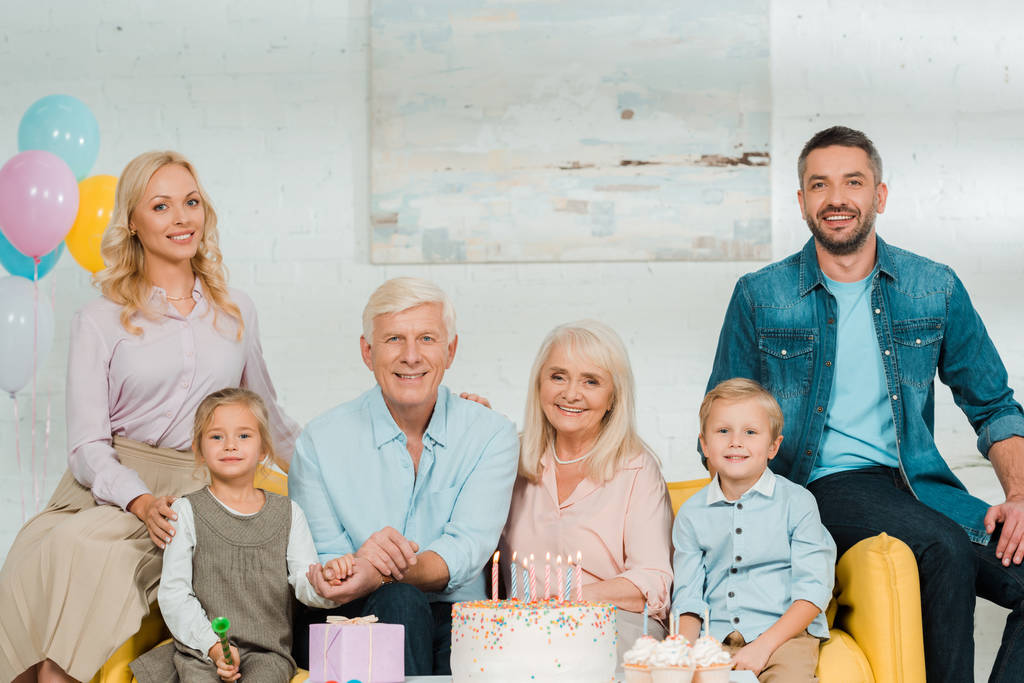 onnellinen perhe istuu lähellä syntymäpäiväkakkua ja katselee kameraa
 - Valokuva, kuva