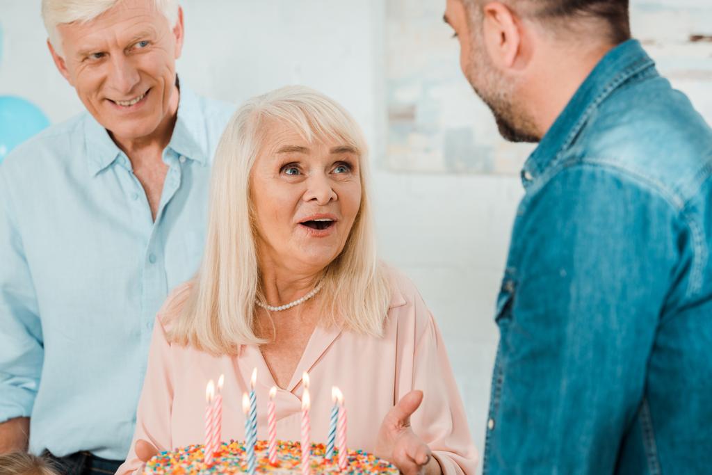 innoissaan vanhempi nainen tilalla syntymäpäivä kakku seisoessaan lähellä aviomies ja aikuinen poika
 - Valokuva, kuva