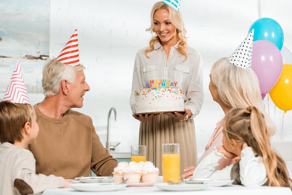 mooie vrouw presenteren verjaardag taart aan senior man zitten aan keukentafel in de buurt van familie - Foto, afbeelding