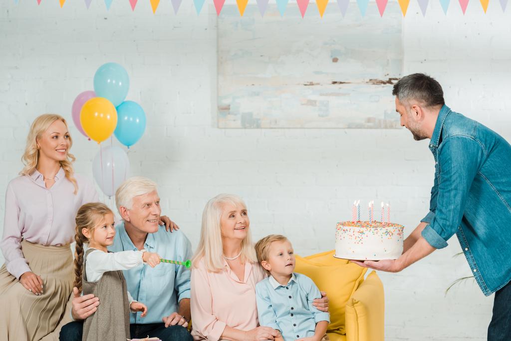 家族の近くのソファに座っている先輩に誕生日ケーキをプレゼントするハンサムな男 - 写真・画像