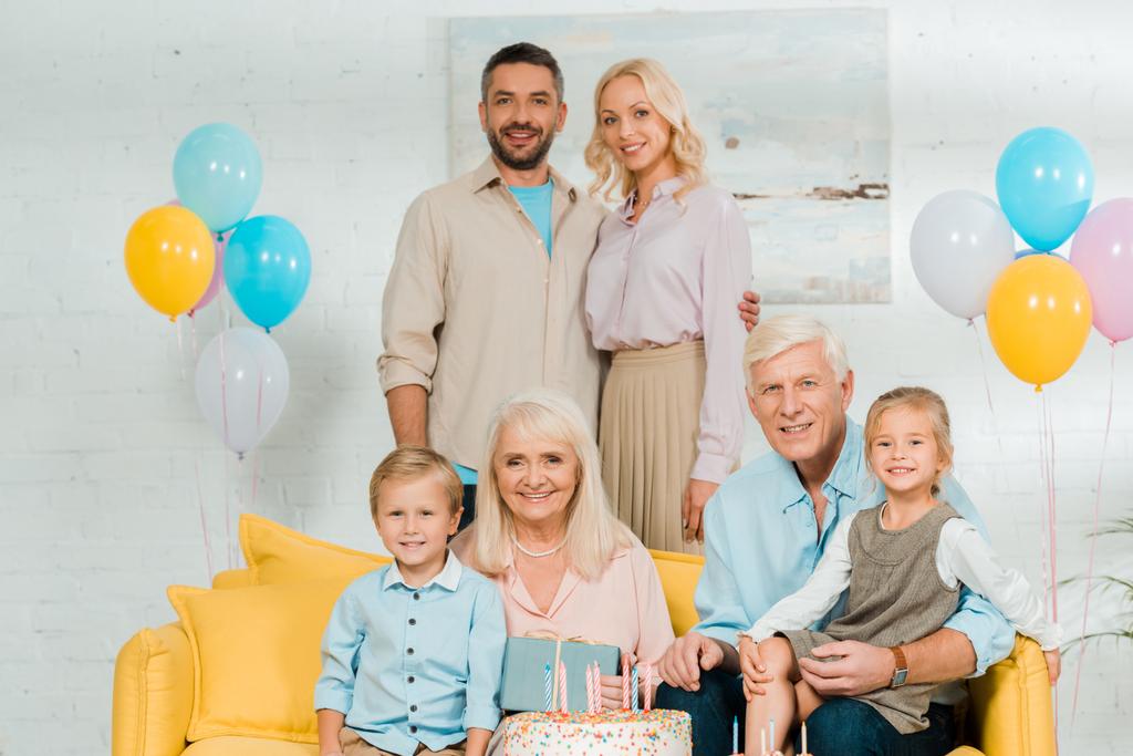 onnellinen perhe katselee kameraa lähellä syntymäpäiväkakkua ja värikkäitä juhlapalloja
 - Valokuva, kuva