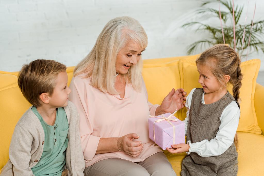 gelukkig senior vrouw nemen geschenk doos van kleindochter terwijl zitten op de gele bank met kleinzoon - Foto, afbeelding