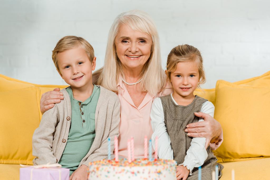 szczęśliwy babcia obejmując wnuki siedząc w pobliżu tort urodzinowy i uśmiechając się do kamery - Zdjęcie, obraz