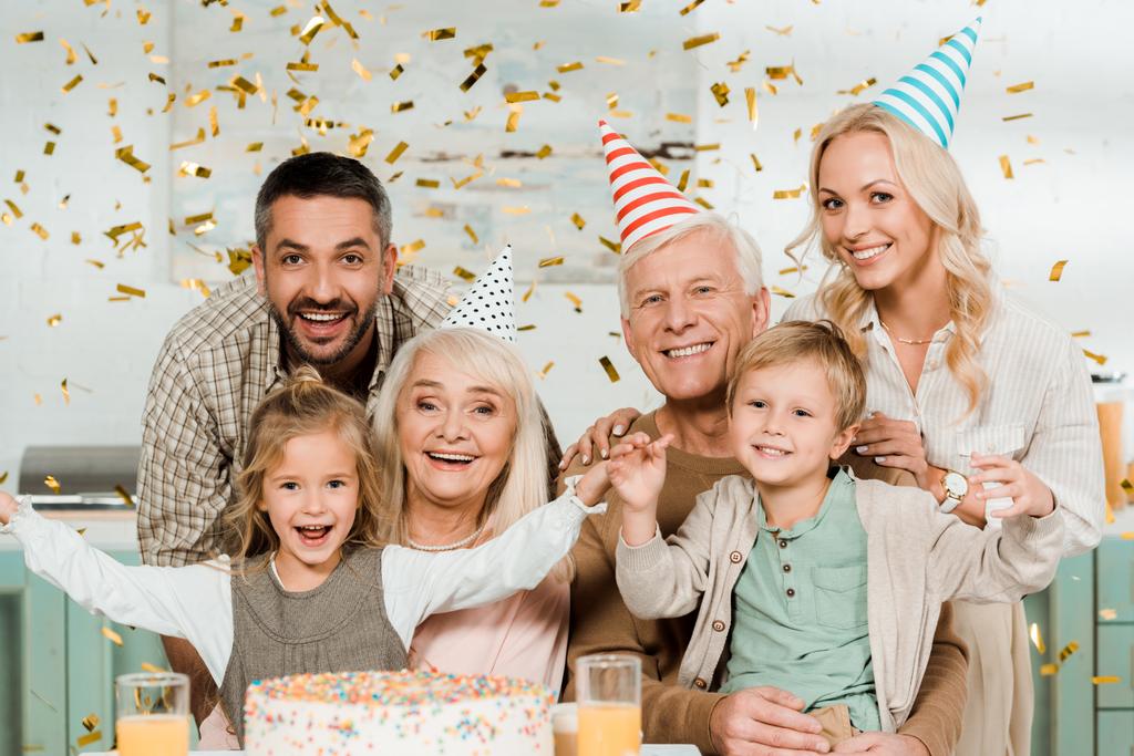 ευτυχισμένη οικογένεια κάθεται κάτω από την πτώση κομφετί κοντά τούρτα γενεθλίων και χαμογελώντας στην κάμερα - Φωτογραφία, εικόνα