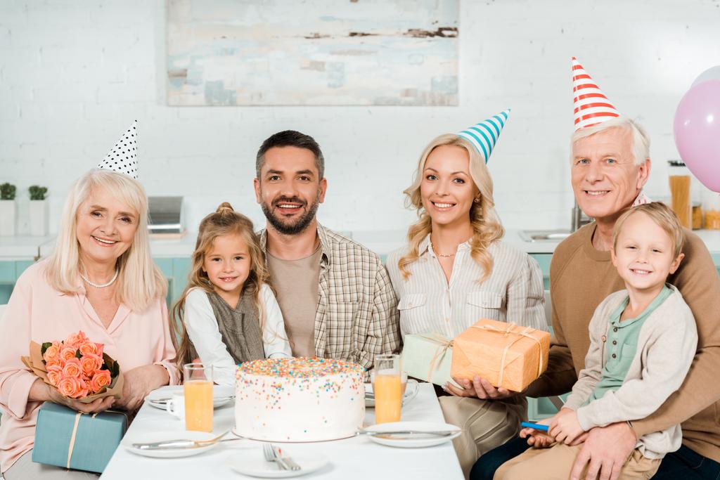 iloinen perhe istuu keittiön pöydässä lähellä syntymäpäiväkakkua ja katselee kameraa
 - Valokuva, kuva