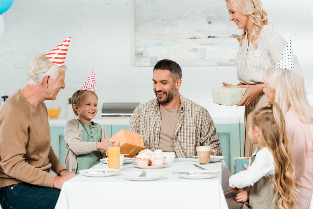 χαμογελαστή γυναίκα και παιδί παρουσιάζει δώρα στον ευτυχισμένο πατέρα κάθεται στο τραπέζι της κουζίνας κοντά στην οικογένεια - Φωτογραφία, εικόνα