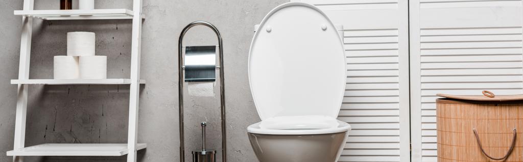 εσωτερικό μοντέρνο μπάνιο με λεκάνη τουαλέτας κοντά rack με πετσέτες, χαρτί υγείας, καλάθι πλυντηρίου και βούρτσα τουαλέτας, πανοραμική λήψη - Φωτογραφία, εικόνα
