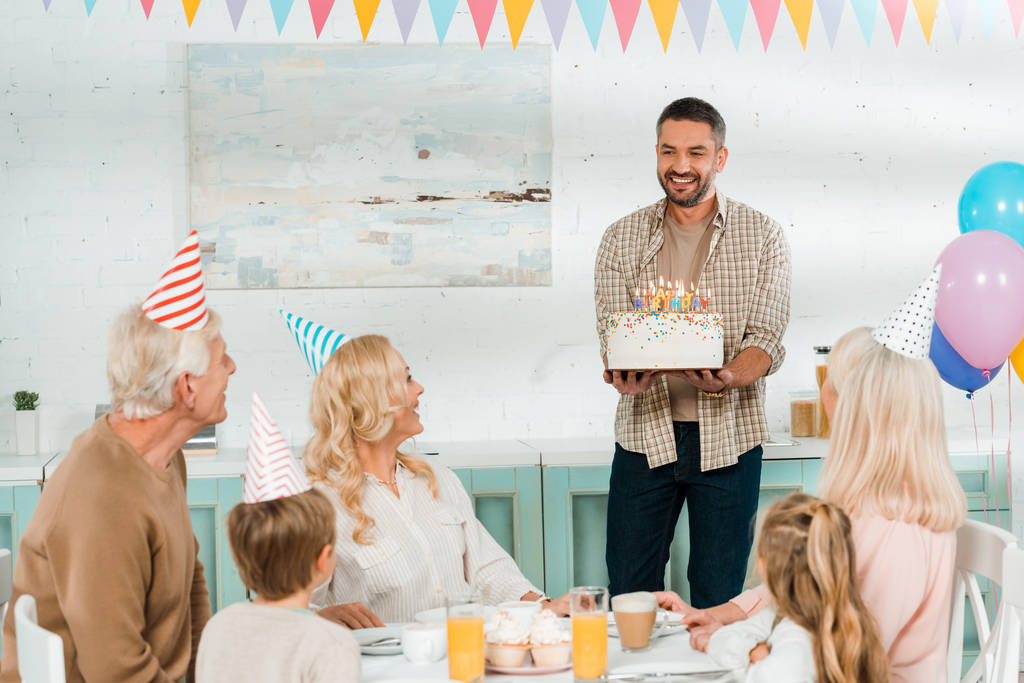 hombre sonriente sosteniendo pastel de cumpleaños con velas cerca de la familia feliz sentado en la mesa de la cocina
 - Foto, Imagen