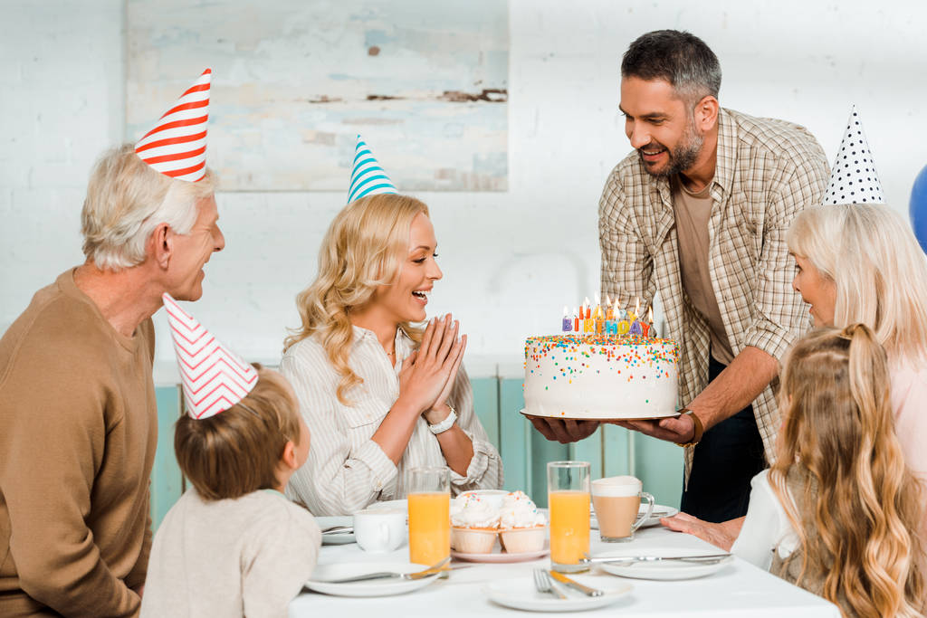 Улыбающийся мужчина держит торт со свечами возле счастливой семьи, сидящей за кухонным столом
 - Фото, изображение